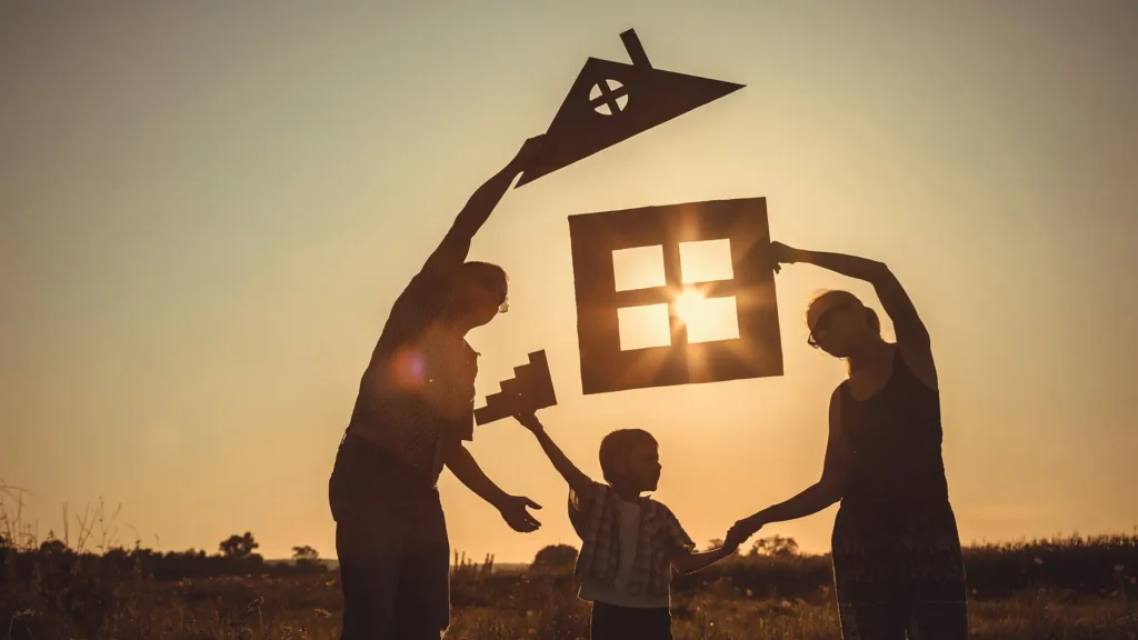 Happy family standing on the field holding up a paper cutout of a house at sunset